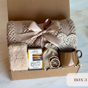 May include: A brown gift box filled with cozy items, including a knitted throw blanket, a mug, cookies, a wooden spoon, and a heart-shaped decoration. The box is labeled "BOX 3".