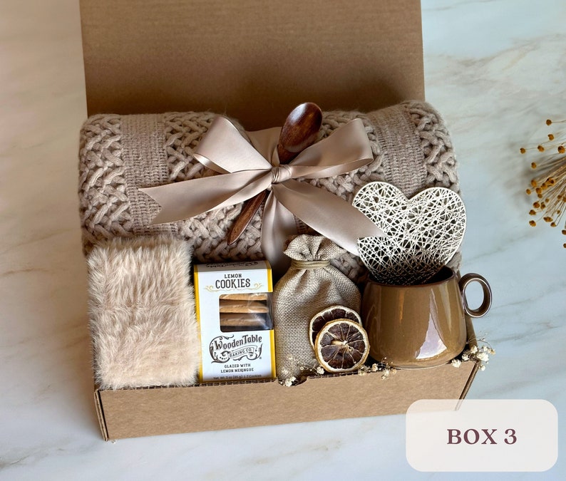 May include: A brown gift box filled with cozy items, including a knitted throw blanket, a mug, cookies, a wooden spoon, and a heart-shaped decoration. The box is labeled "BOX 3".