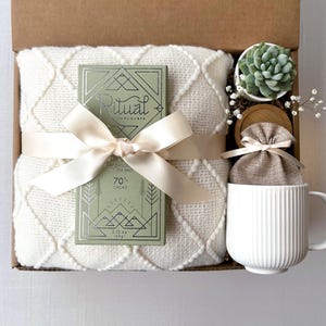 K&ouml;nnte beinhalten: Eine Geschenkbox gef&uuml;llt mit gem&uuml;tlichen Artikeln, darunter eine wei&szlig;e Decke, eine Tafel Ritual-Schokolade, eine kleine Sukkulente im Topf, eine wei&szlig;e Tasse und ein kleiner Jutesack. Die Schokoladentafel ist in gr&uuml;nes Papier eingewickelt, auf dem die Worte "Ritual Chocolate" in Silber gedruckt sind.
