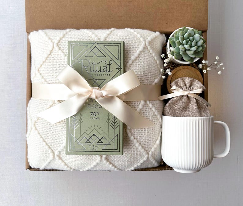 K&ouml;nnte beinhalten: Eine Geschenkbox gef&uuml;llt mit gem&uuml;tlichen Artikeln, darunter eine wei&szlig;e Decke, eine Tafel Ritual-Schokolade, eine kleine Sukkulente im Topf, eine wei&szlig;e Tasse und ein kleiner Jutesack. Die Schokoladentafel ist in gr&uuml;nes Papier eingewickelt, auf dem die Worte "Ritual Chocolate" in Silber gedruckt sind.