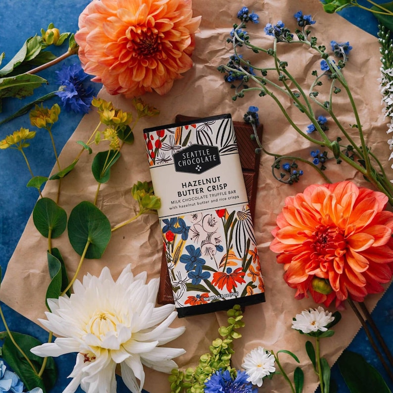 Puede incluir: Una barra de trufa de chocolate con leche con mantequilla de avellanas y arroz crujiente, rodeada de flores coloridas. El empaque presenta el texto "HAZELNUT BUTTER CRISP" y "SEATTLE CHOCOLATE". Las flores incluyen dalias naranjas, acianos azules y dalias blancas.
