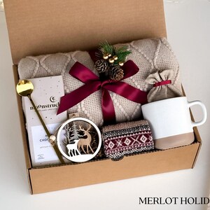 Puede incluir: Una caja de regalo con una manta de punto trenzado color crema, una taza blanca con base beige, una cuchara dorada, una tableta de chocolate, un adorno de ciervo de madera y calcetines estampados. La caja est&aacute; atada con una cinta burdeos y contiene el texto "MERLOT HOLIDAY".