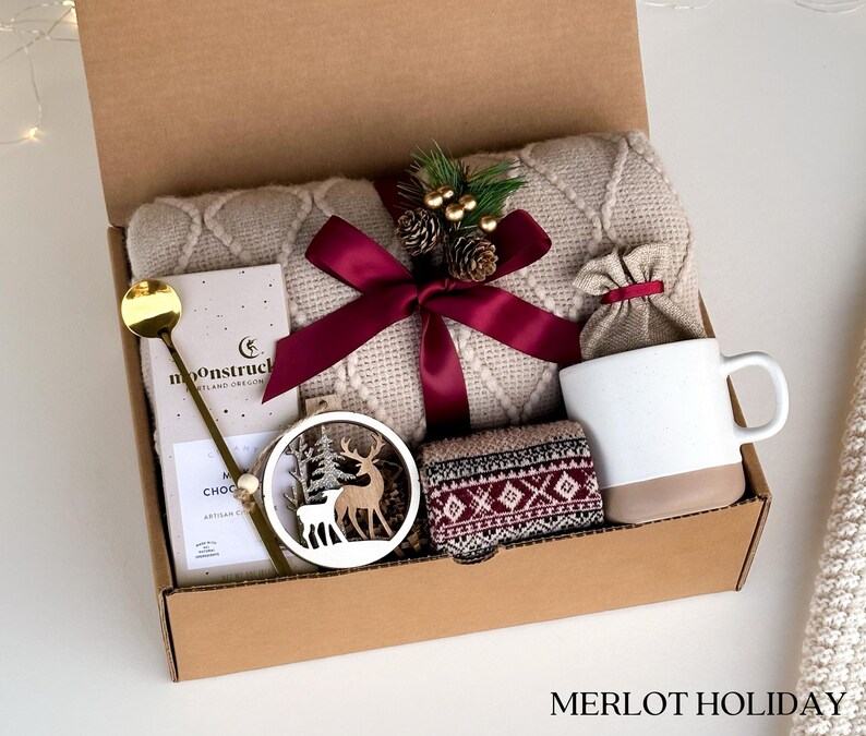 Puede incluir: Una caja de regalo con una manta de punto trenzado color crema, una taza blanca con base beige, una cuchara dorada, una tableta de chocolate, un adorno de ciervo de madera y calcetines estampados. La caja est&aacute; atada con una cinta burdeos y contiene el texto "MERLOT HOLIDAY".