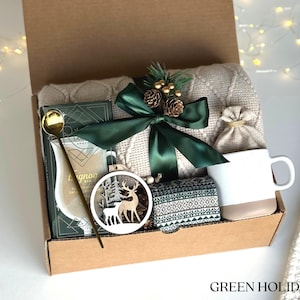May include: A brown cardboard gift box filled with a cozy winter-themed gift set. The box contains a white blanket, a mug, a small bag, a wooden ornament with a deer design, a gold spoon, and a box of eggnog mix. The box is tied with a green ribbon and decorated with a small pine cone and berries.