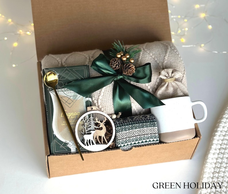 May include: A brown cardboard gift box filled with a cozy winter-themed gift set. The box contains a white blanket, a mug, a small bag, a wooden ornament with a deer design, a gold spoon, and a box of eggnog mix. The box is tied with a green ribbon and decorated with a small pine cone and berries.