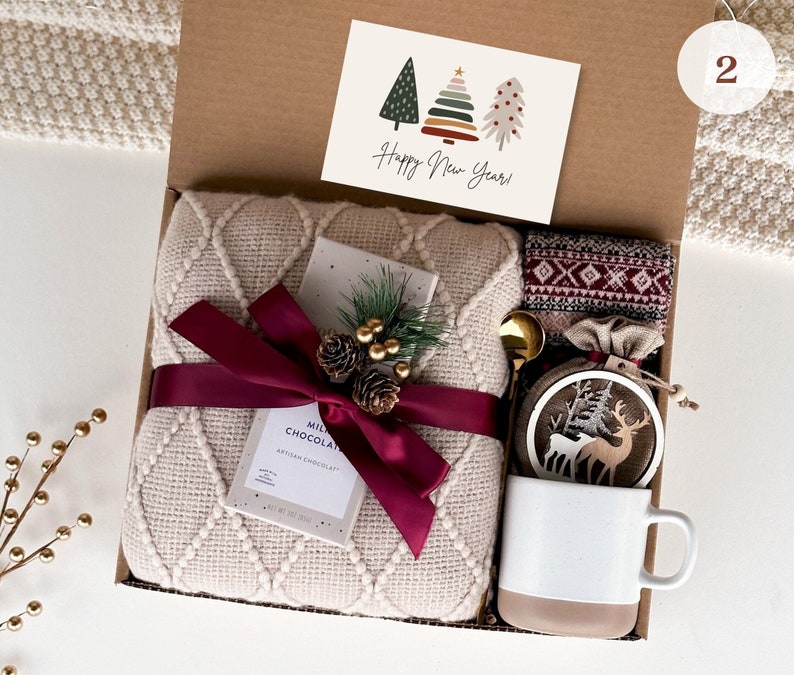 May include: A gift box filled with cozy winter items, including a cream-colored blanket, a red and white patterned scarf, a mug, a hot chocolate packet, a wooden ornament with a deer design, and a card that says "Happy New Year!"