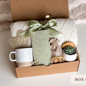 May include: A brown cardboard box filled with a variety of gifts, including a white mug, a small potted succulent, a bag of bath salts, a white blanket, a gold heart-shaped ornament, and a green and white box of bath salts. The box is labeled "BOX 4".