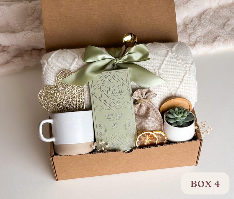 May include: A brown cardboard box filled with a variety of gifts, including a white mug, a small potted succulent, a bag of bath salts, a white blanket, a gold heart-shaped ornament, and a green and white box of bath salts. The box is labeled "BOX 4".