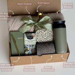 May include: A gift box filled with cozy items, including a soft blanket, a heart-shaped coaster, a gold spoon, a stainless steel travel tumbler, a vegan leather notebook with a gold pen, gourmet chocolate, and warm wool socks. The box also includes a gift box greeting card and ribbon.