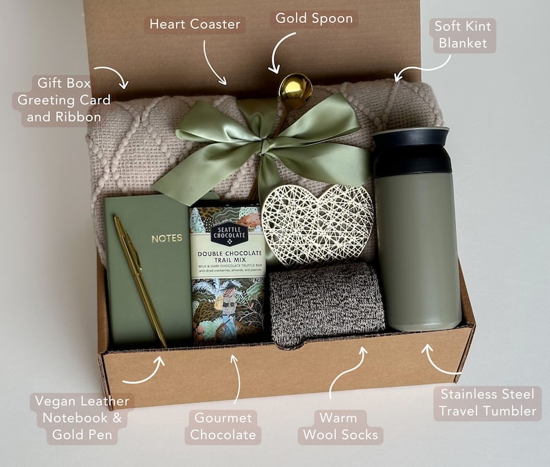 May include: A gift box filled with cozy items, including a soft blanket, a heart-shaped coaster, a gold spoon, a stainless steel travel tumbler, a vegan leather notebook with a gold pen, gourmet chocolate, and warm wool socks. The box also includes a gift box greeting card and ribbon.