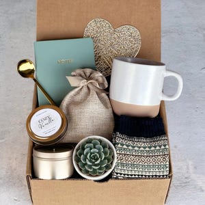 K&ouml;nnte beinhalten: Eine Geschenkbox gef&uuml;llt mit gem&uuml;tlichen Artikeln, darunter eine Tasse, eine Kerze, ein Notizbuch, eine kleine Pflanze und Socken. Die Box ist braun und hat eine goldene herzf&ouml;rmige Dekoration auf dem Deckel.