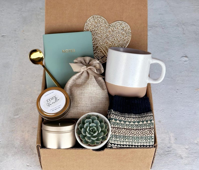 K&ouml;nnte beinhalten: Eine Geschenkbox gef&uuml;llt mit gem&uuml;tlichen Artikeln, darunter eine Tasse, eine Kerze, ein Notizbuch, eine kleine Pflanze und Socken. Die Box ist braun und hat eine goldene herzf&ouml;rmige Dekoration auf dem Deckel.