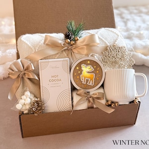 May include: A brown cardboard gift box filled with a white blanket, a mug, a bag of marshmallows, a box of hot cocoa mix, and a wooden reindeer ornament. The box is decorated with a gold ribbon and a snowflake.