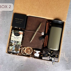 May include: A brown cardboard box with the text "BOX 2" on the front. The box is open and contains a variety of items, including a brown leather journal, a gold pen, a scented candle, a chocolate bar, a travel mug, and a small jar of jam. The items are arranged on a bed of brown shredded paper.