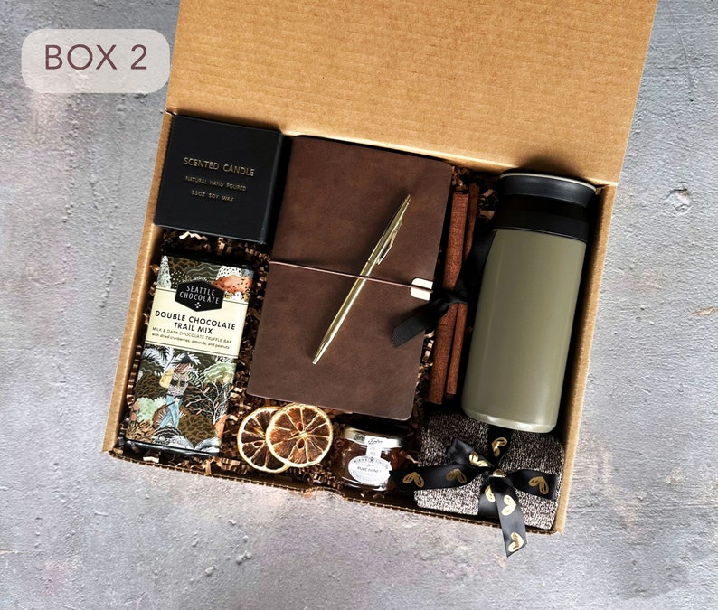May include: A brown cardboard box with the text "BOX 2" on the front. The box is open and contains a variety of items, including a brown leather journal, a gold pen, a scented candle, a chocolate bar, a travel mug, and a small jar of jam. The items are arranged on a bed of brown shredded paper.