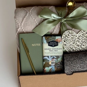 May include: A gift box filled with items including a blanket, a notebook with a gold pen, a chocolate bar with the text "Seattle Chocolate Double Chocolate Trail Mix", a heart-shaped decorative object, and a pair of socks.