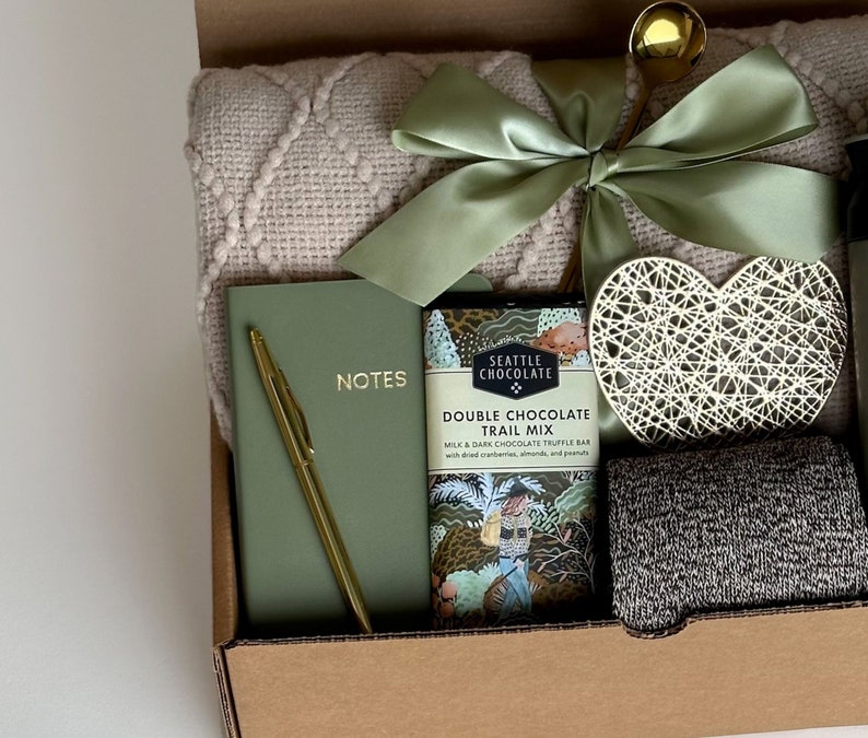 May include: A gift box filled with items including a blanket, a notebook with a gold pen, a chocolate bar with the text "Seattle Chocolate Double Chocolate Trail Mix", a heart-shaped decorative object, and a pair of socks.