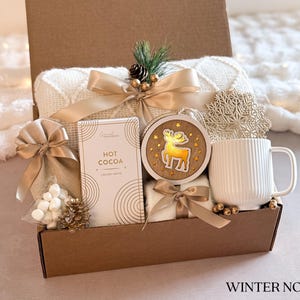 Op de afbeelding: Een bruine kartonnen cadeaudoos gevuld met een wit deken, een mok, een zak marshmallows, een doos met warme chocolademelk en een houten rendier ornament. De doos is versierd met een gouden lint en een sneeuwvlok.