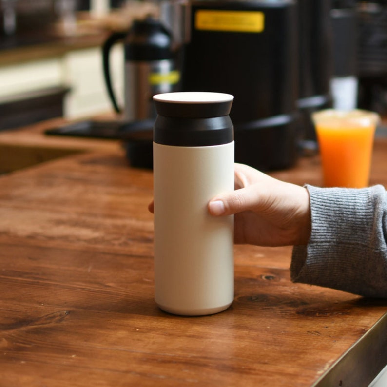 Puede incluir: Un termo de viaje aislado de color crema con tapa y borde negros. El termo cil&iacute;ndrico es sostenido por una mano sobre una mesa de madera. El fondo incluye una cafetera y un vaso de zumo de naranja.