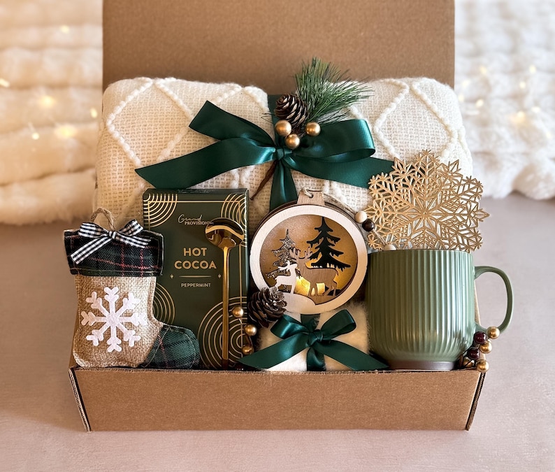 May include: A brown gift box filled with a cozy white blanket, a green mug, a wooden ornament with a deer and pine trees, a gold snowflake stocking, a box of hot cocoa mix, and a green ribbon.