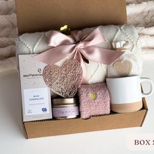 May include: A gift box filled with cozy items, including a white blanket, a mug, a candle, a bar of chocolate, and a pair of pink socks. The box is decorated with a pink ribbon and a heart-shaped ornament. The text on the chocolate bar reads "Milk Chocolate Artisan Chocolate". The text on the candle reads "Hug in a Tin".