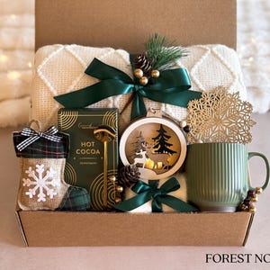 May include: A gift basket with a white blanket, a green mug, a wooden ornament with a deer and a Christmas tree, a box of hot cocoa mix, a gold spoon, and a green and plaid stocking with a snowflake.