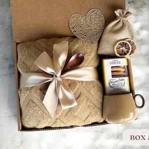 May include: A gift box filled with a brown blanket, a wooden spoon, a bag of cookies, a mug, and a heart-shaped coaster. The box is tied with a beige ribbon and the cookies are labeled "Lemon Cookies".
