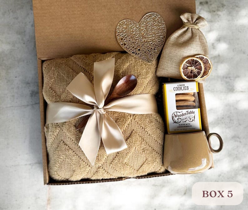 May include: A gift box filled with a brown blanket, a wooden spoon, a bag of cookies, a mug, and a heart-shaped coaster. The box is tied with a beige ribbon and the cookies are labeled "Lemon Cookies".