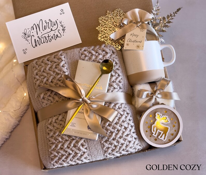 May include: A gift box filled with cozy items for the winter. It includes a knitted throw blanket, a mug, a gold spoon, a bar of chocolate, a small gift bag, and a wooden moose ornament. The box is decorated with a gold ribbon and a card that says "Merry Christmas".