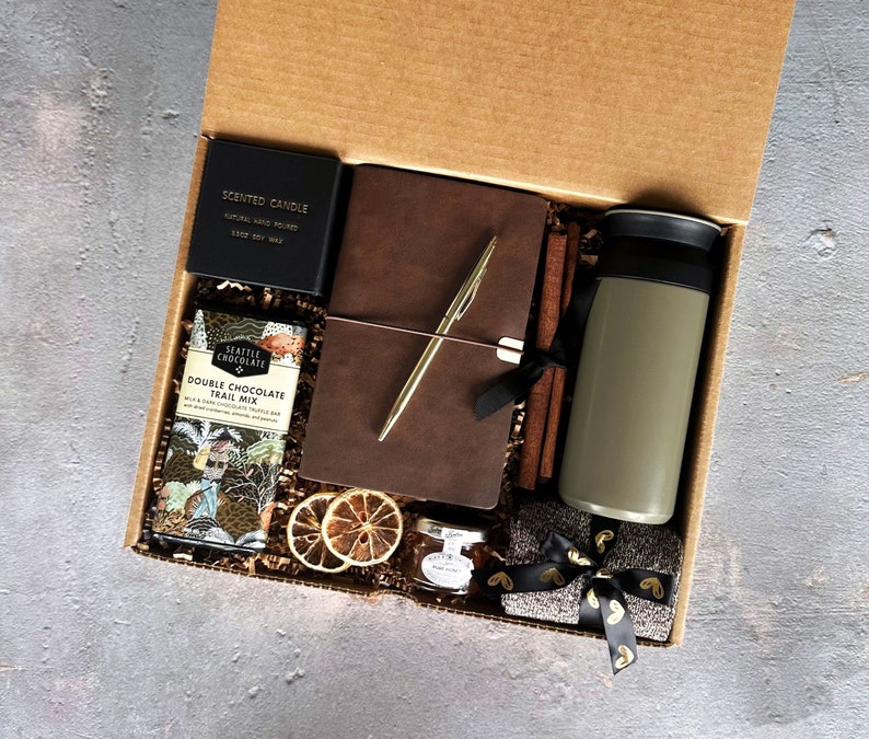 May include: A brown gift box filled with a variety of items, including a brown leather journal, a gold pen, a scented candle, a jar of jam, a bag of chocolate trail mix, a travel mug, and a grey scarf with gold ribbon.