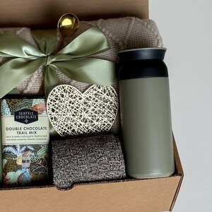 May include: A gift basket with a green and white blanket, a green insulated water bottle, a heart-shaped decorative piece, a chocolate bar with the text "Seattle Chocolate Double Chocolate Trail Mix", and a gray knitted scarf.