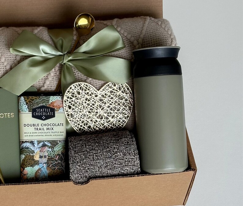 May include: A gift basket with a green and white blanket, a green insulated water bottle, a heart-shaped decorative piece, a chocolate bar with the text "Seattle Chocolate Double Chocolate Trail Mix", and a gray knitted scarf.