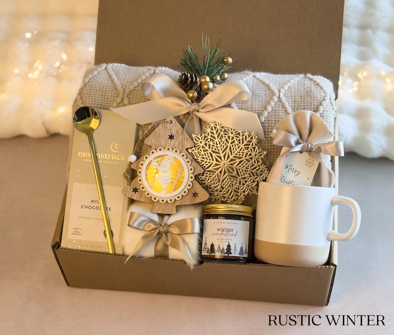 Op de afbeelding: Een rustieke wintercadeaudoos gevuld met een knusse deken, een mok, een kaars, warme chocolademelk en een houten kerstboomversiering. De doos is versierd met een gouden sneeuwvlok en een label met de tekst "Merry Christmas".