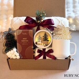 Puede incluir: Una caja de regalo navide&ntilde;a con una manta blanca, chocolate caliente, una taza blanca, un adorno con ciervos y &aacute;rboles, un adorno de &aacute;rbol de Navidad a cuadros y malvaviscos. La caja est&aacute; atada con una cinta burdeos y decorada con detalles dorados.