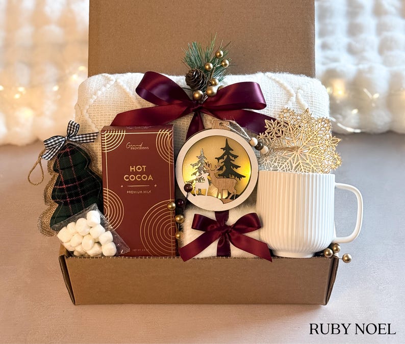 Puede incluir: Una caja de regalo navide&ntilde;a con una manta blanca, chocolate caliente, una taza blanca, un adorno con ciervos y &aacute;rboles, un adorno de &aacute;rbol de Navidad a cuadros y malvaviscos. La caja est&aacute; atada con una cinta burdeos y decorada con detalles dorados.