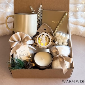 Puede incluir: Una caja de regalo navide&ntilde;a llena de una taza beige, un cuaderno, un bol&iacute;grafo, un adorno de &aacute;rbol de Navidad de madera, una vela, una bolsa de malvaviscos y un peque&ntilde;o art&iacute;culo esponjoso, todos atados con cintas. La caja tiene las palabras "Warm Wishes" impresas.