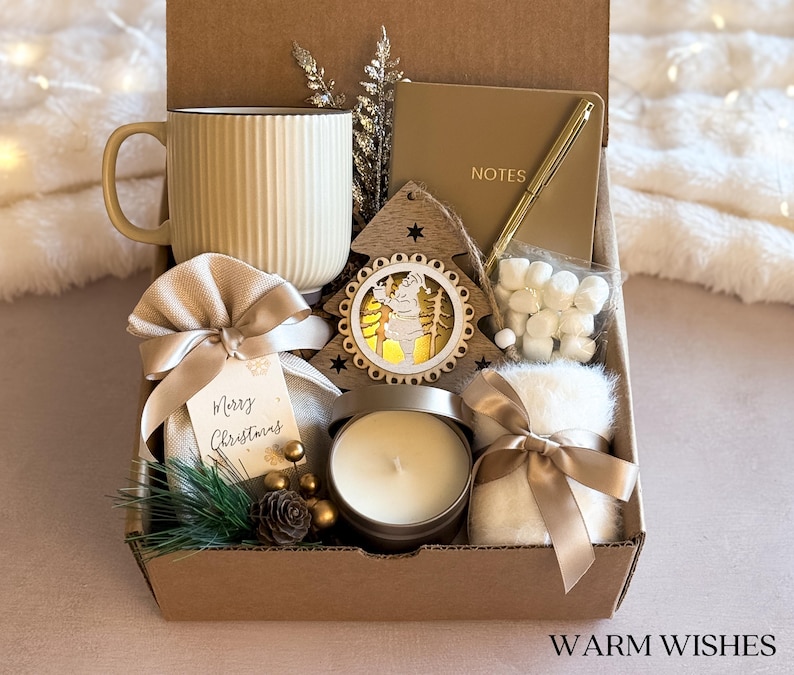 Puede incluir: Una caja de regalo navide&ntilde;a llena de una taza beige, un cuaderno, un bol&iacute;grafo, un adorno de &aacute;rbol de Navidad de madera, una vela, una bolsa de malvaviscos y un peque&ntilde;o art&iacute;culo esponjoso, todos atados con cintas. La caja tiene las palabras "Warm Wishes" impresas.