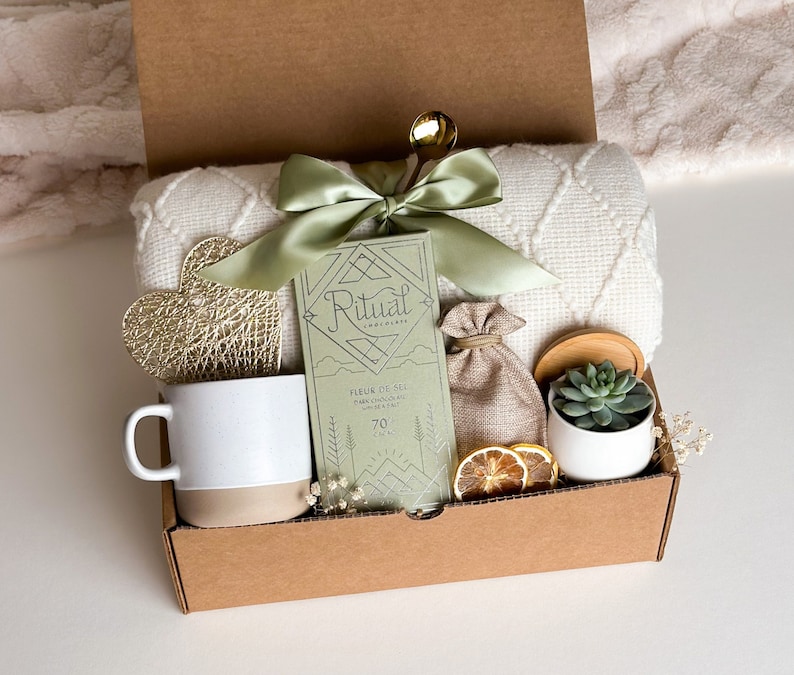 May include: A brown gift box filled with a variety of items, including a white mug, a bar of chocolate, a small plant, a decorative heart, and a white throw blanket. The box is tied with a green ribbon and has the word "Ritual" printed on the chocolate bar.