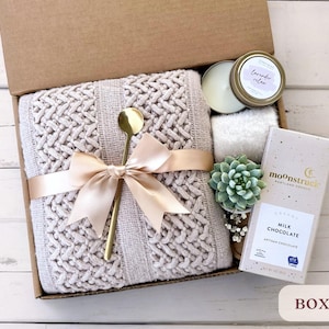 May include: A gift box filled with self-care items, including a knitted throw blanket, a candle, a succulent plant, and a bar of chocolate. The box is tied with a satin ribbon and labeled "Box 7".