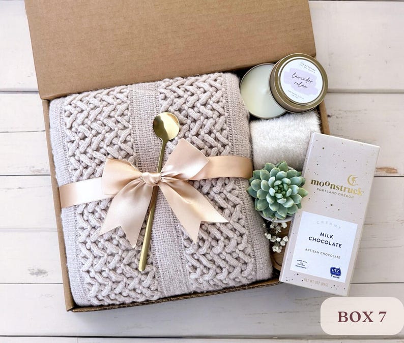 May include: A gift box filled with self-care items, including a knitted throw blanket, a candle, a succulent plant, and a bar of chocolate. The box is tied with a satin ribbon and labeled "Box 7".