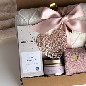 Peut inclure: Une bo&icirc;te-cadeau remplie d'articles de soins personnels, dont une couverture, une d&eacute;coration en m&eacute;tal en forme de c&oelig;ur, une bougie, une barre de chocolat et une paire de chaussettes en fourrure. La bo&icirc;te est d&eacute;cor&eacute;e d'un ruban rose et d'un c&oelig;ur dor&eacute;.