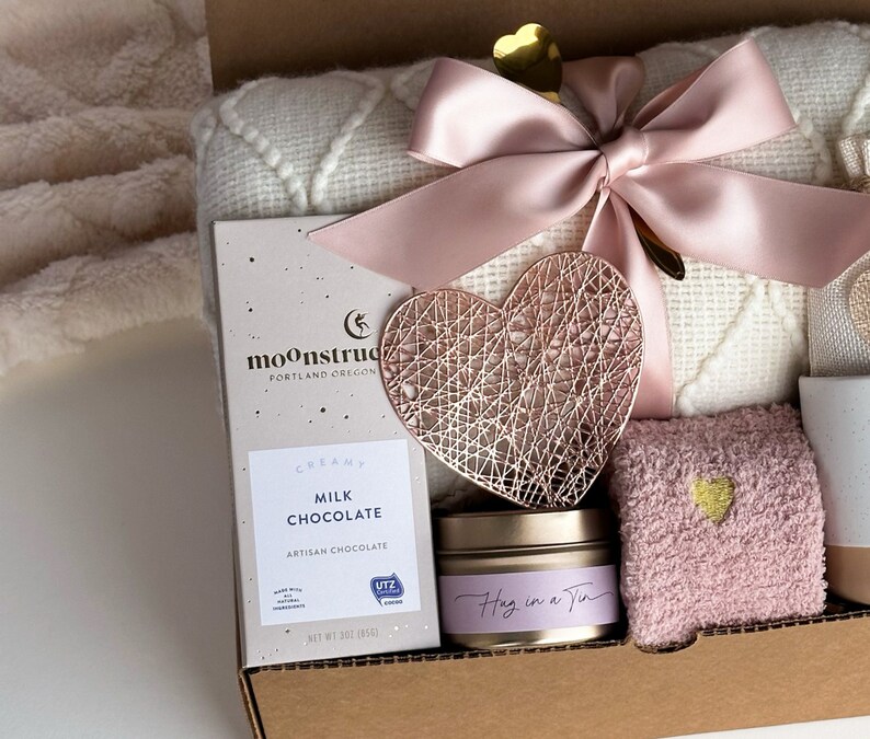 Peut inclure: Une bo&icirc;te-cadeau remplie d'articles de soins personnels, dont une couverture, une d&eacute;coration en m&eacute;tal en forme de c&oelig;ur, une bougie, une barre de chocolat et une paire de chaussettes en fourrure. La bo&icirc;te est d&eacute;cor&eacute;e d'un ruban rose et d'un c&oelig;ur dor&eacute;.