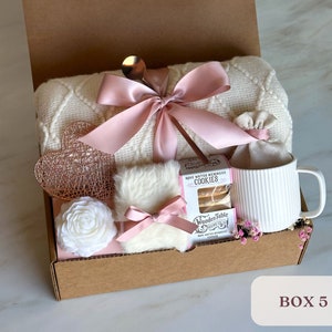 May include: A gift box filled with a white blanket, a white mug, a white faux fur, a white rose, a rose water meringue cookie, and a pink ribbon. The box is labeled "BOX 5".