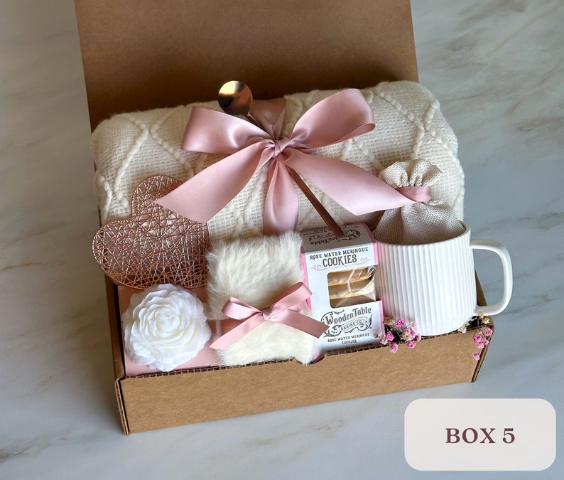 May include: A gift box filled with a white blanket, a white mug, a white faux fur, a white rose, a rose water meringue cookie, and a pink ribbon. The box is labeled "BOX 5".