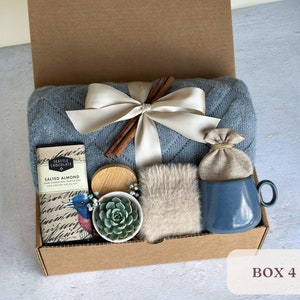 May include: A blue and white gift box filled with a variety of items, including a blue mug, a small plant, a brown bag, a chocolate bar, and a piece of fabric. The box is labeled "Box 4".