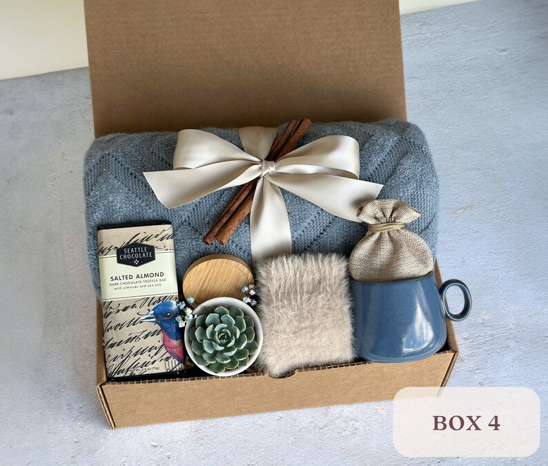 May include: A blue and white gift box filled with a variety of items, including a blue mug, a small plant, a brown bag, a chocolate bar, and a piece of fabric. The box is labeled "Box 4".