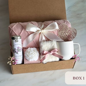 May include: A pink and white gift box filled with a pink knitted throw blanket, a white mug, a white candle, a small white fluffy pillow with a pink ribbon, a tin of tea, and a decorative heart. The box is labeled "BOX 1".