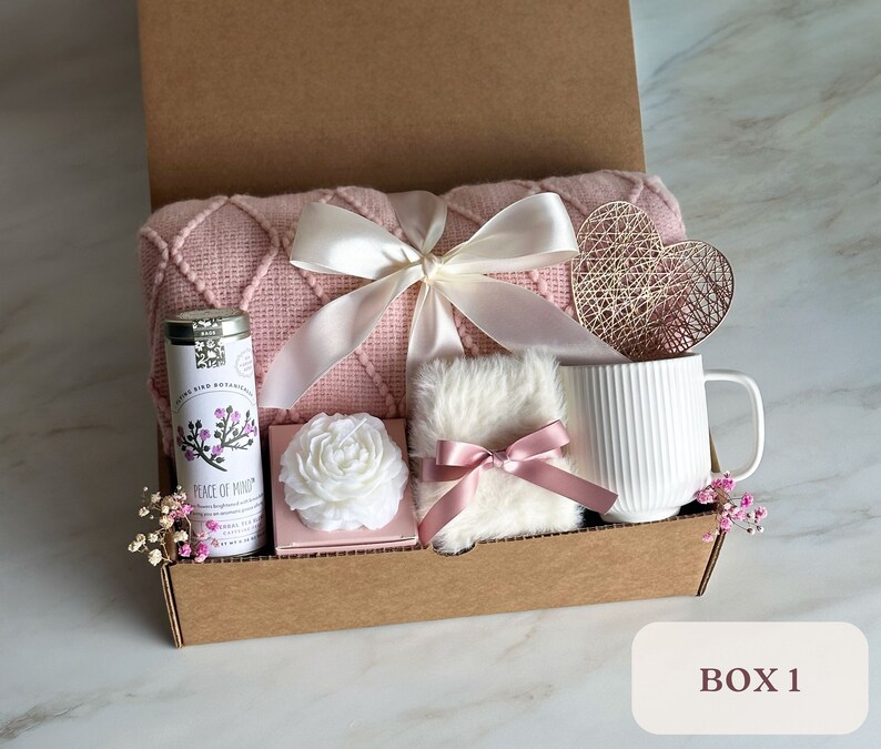 May include: A pink and white gift box filled with a pink knitted throw blanket, a white mug, a white candle, a small white fluffy pillow with a pink ribbon, a tin of tea, and a decorative heart. The box is labeled "BOX 1".