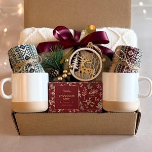 May include: A gift basket filled with cozy items for the winter holidays. It includes a white blanket, two pairs of patterned socks, two mugs, a box of chocolate chip cookies, and a gold reindeer ornament.