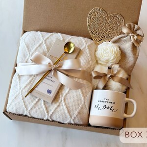 May include: A gift box filled with a white blanket, a bar of milk chocolate, a gold spoon, a white flower, a small bag, a heart-shaped gold ornament, and a white mug with the text "The World's Best Mom" in black. The box is labeled "Box 7".